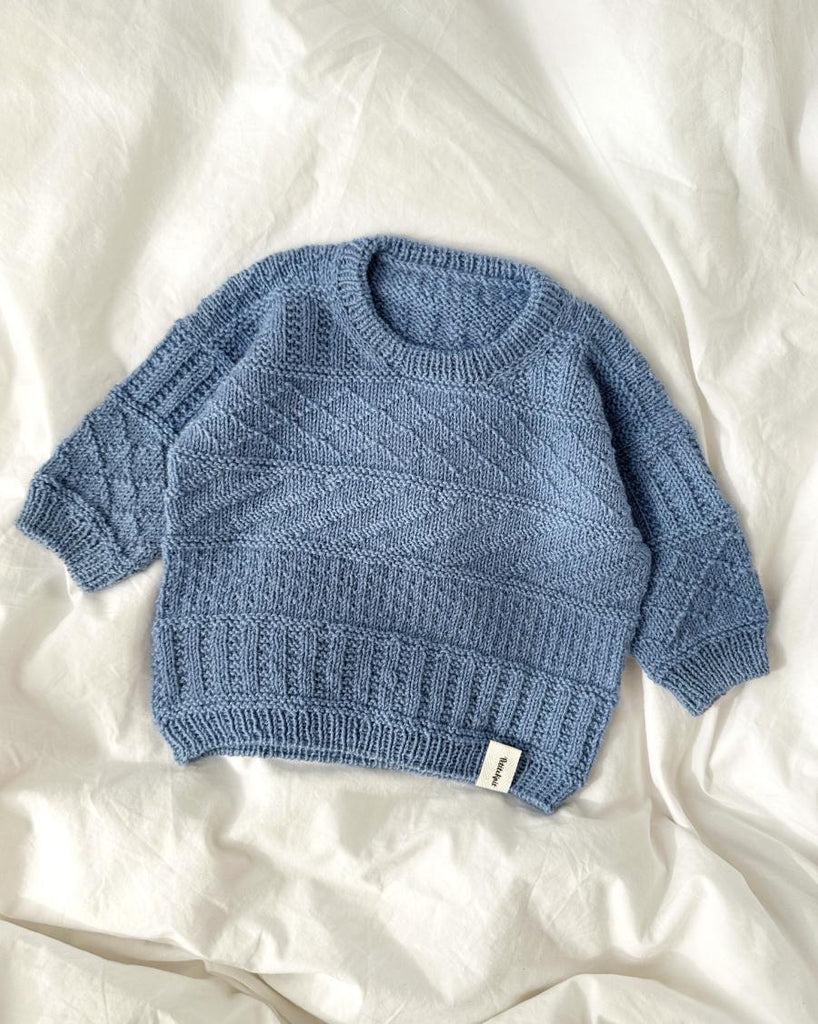 Lyseblå Storm Sweater Baby liggende på sengetøj